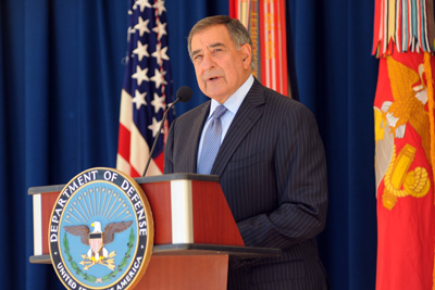 A man in a suit stands behind a lectern, which bears the seal of the Department of Defense. Behind him are several flags, including that of the United States and the Marine Corps.