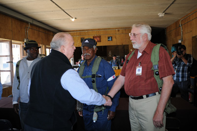 Inside a small room, two civilian men shake hands.  Other men stand nearby.