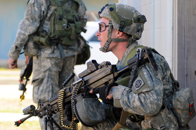 A kneeling soldier holds an automatic, belt-fed weapon, Another soldier stands nearby.