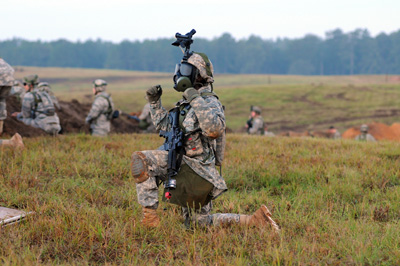 In the middle of a grassy field, a soldier is down on one knee. He wears a gas mask and an optical device on his helmet. Other soldiers are nearby.  