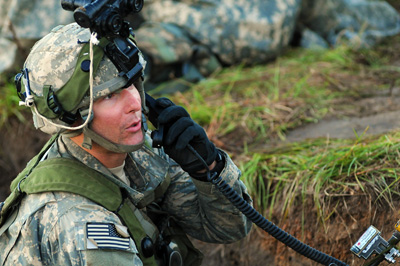 A man in a military uniform in a grassy and muddy environment talks on a mobile radio.  He has optical gear attached to his combat helmet.  In the rear, the uniforms of other soldiers can be seen.  