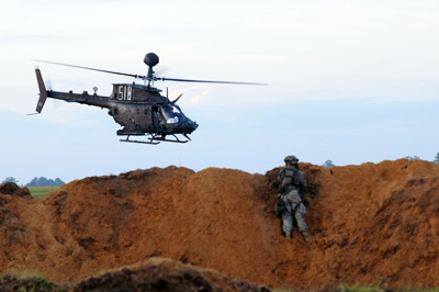 A single Soldier lays prone on a large dirt berm.  Overhead he sees a helicopter in the air. The helicopter has the numbers "518" painted on the side.