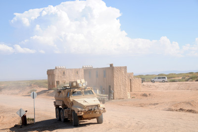 In a desert environment a military combat vehicle kicks up dust as it rolls down a road.  In the read is a concrete building.  A huge cloud hangs in an otherwise bright blue sky.
