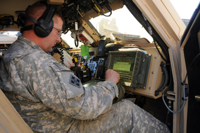 A Soldier sits in a combat vehicle and operated a computer device. He wears headphones.
