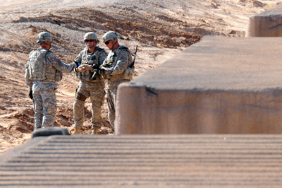 Near three large concrete blocks stand three Soldiers in a desert environment.