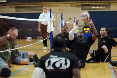 In the middle of a gymnasium, multiple people are engaged in a game of volleyball but all players are seated on the ground. One player has hit the ball into the air.