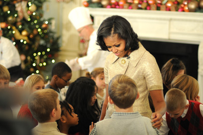 A woman is surrounded by small children. In the rear is a fireplace, a Christmas tree, and a man in a chef's outfit.