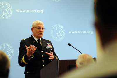 A man in a military uniform stands behind a lectern.  Behind him the wall has the words "www.usaa.org" printed in multiple places. People in the foreground watch him.