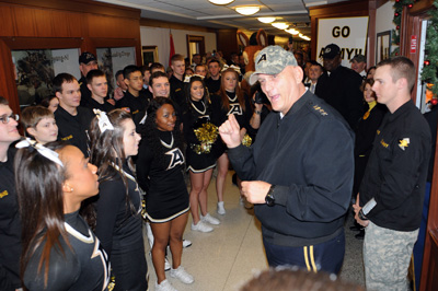 A man in a military uniform and a camouflage baseball cap with an "A" on it talks to a group of young people in sportswear. A sign in the background says "Go Army!"