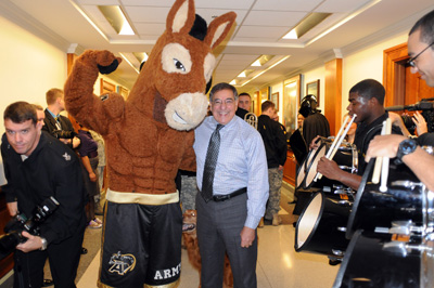 In a large hallway, a person in a mule mascot costume wears shorts that say "Army."  He stands next to a man in a blue shirt with a tie.  Men playing drums are nearby.