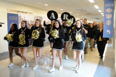 Cheerleaders walk down a large hallway.  Dozens of people follow.  Some carry cardboard circles that spell out the word "Army."