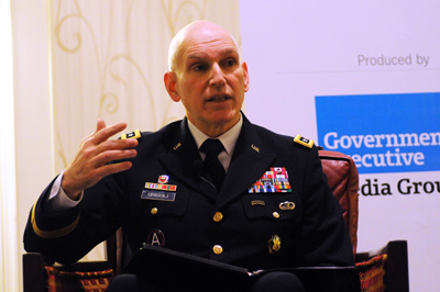 A man in a military uniform sits in a leather chair. His hand is held up int he air.  Behind him is a sign that says "Powered by Government Executive Media Group."