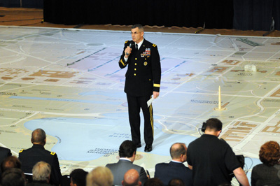 A man in a military uniform stands in the center of a huge map that covers the floor of a large room.  In front of him are a dozen people listening to him speak.