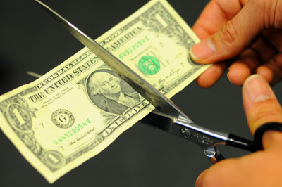 A hand holds a dollar bill. Another hand holds a pair of scissors and appears to cut the bill.