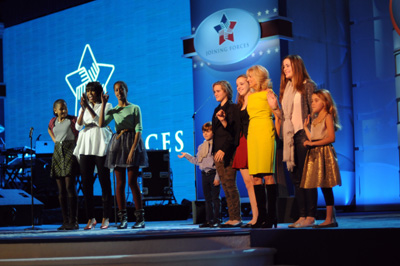 A woman and several children stand together on a stage.  Nearby are three other women standing together.  A sign hanging at the back of the stage reads "Joining Forces."
