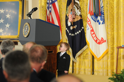 A small boy stands behind a lectern that is two feet taller than him. He explores the back side of the lectern.  Behind him are several flags, including the U.S. flag.
