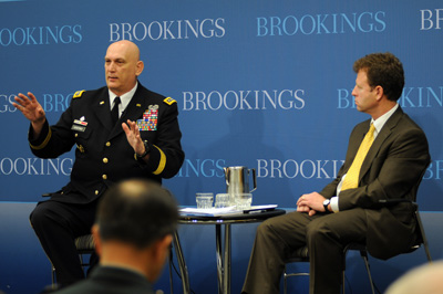 A man in a military uniform and a man in a suit site across from each other at a tiny table.  The uniformed man is speaking to people off camera.  Behind both men is a wall that has the words "Brookings" printed on it multiple times,
