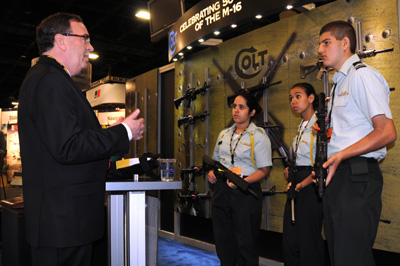 Three young people in military uniforms, all three with rifles, stand in front of a display. A man in a suit speaks with them.