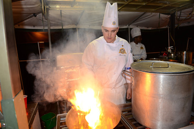 A young man in a chef's outfit tends to a pan that is aflame. Next to him is a large pot.