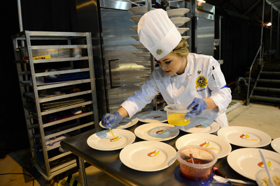 A young woman in a chef's outfit decorates desert plates by drawing red and yellow petals on them with fruit coulis.
