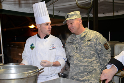 A young man in a chef's hat and a man in a military uniform stand together in front of a large pot.