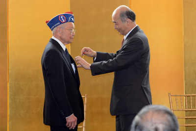 A man in a suit places a medal on the lapel of an elderly man who wears a hat that indicates veteran status.