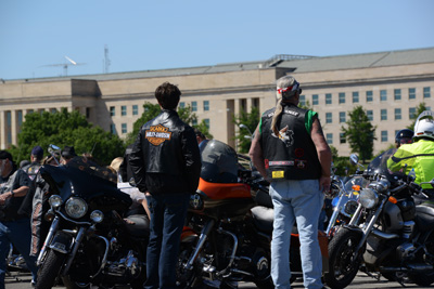 Two men in leather vests that say "Harley Davidson" stand near a row of motorcycles in front of the Pentagon.