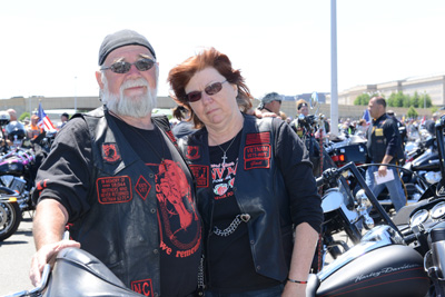 A man and woman, both in leather vests, stand behind a motorcycle.  Dozens of other motorcycles are in the area.