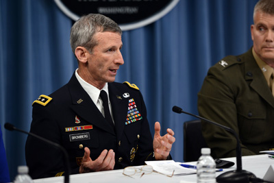 Two men in military uniforms sit at a table. One is talking to another person who is off camera.