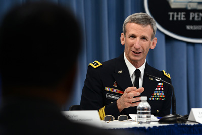 A man in a military uniform sits at a table and speaks with a person in the foreground.