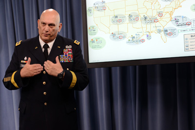 A man in a military uniform stands in front of a television screen with a map of the U.S. on it.