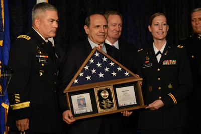 A man and a woman, both in military uniforms, flank another man in a suit who is holding a shadow box with an American flag inside.