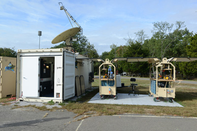 A cargo container with its doors open sits outdoors on a concrete slab.  A satellite dish sits on top of it.  Next to it are two large tool carts. 