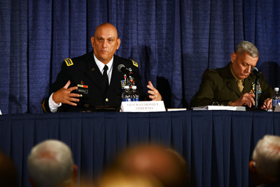 A man in a military uniform sits at a long table with another man in a military uniform.  He speaks to people seated in front of him.