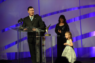 A man in a suit stands behind a lectern.  Near him is a trophy on a pedestal. A woman and small girl stand nearby.