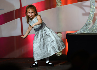 A young girls in a sparkly grey dress with flower outlines dances on a stage.