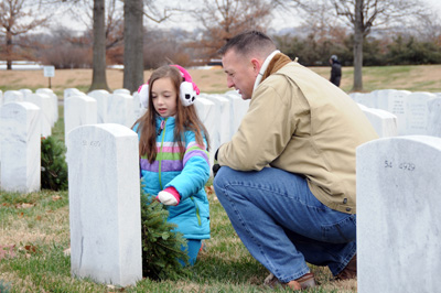 Amidst more than a dozen white gravestones, a young girl places a wreath.  An older man kneels next to her.