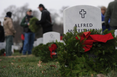 Several white grave markers are adorned with wreaths made of greenery and red ribbons.  The nearest marker says "Alfred" and features a cross.  People stand together in the background.
