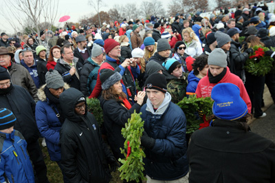 Dozens of people in winter clothing stand together outside.  Many of them carry wreaths made of greenery.