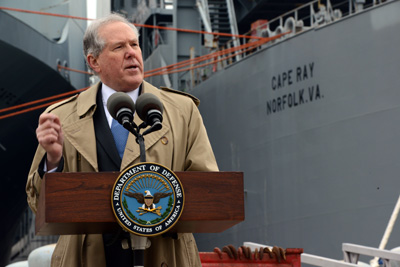 A man stands behind a lectern, which has a Department of Defense insignia on it. Behind him is an ocean-going vessel with the words "Cape Ray Norfolk, Va." printed on its side.