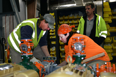 A man and women make adjustments to a piece of colorful industrial equipment. Another man stands nearby and watches.
