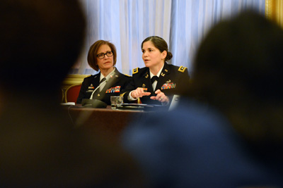 Two women in military uniforms are seated at a table. They are framed by the bodies of people in the foreground.