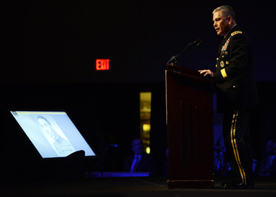 A man in a military uniform stands behind a lectern in a darkened room.  In front of him is a television screen, on the stage, with a photo of a young Soldier. An exit sign appears in the background.