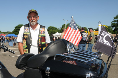 A man wears a leather vest with many patches. He stands outdoors behind a motorcycle that has an American flag and a POW/MIA flag.