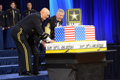 Two men in military uniforms use a sword to cut a cake, which is decorated with American flags and a logo that says "U.S. Army."