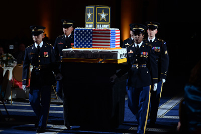 Four individuals in military uniforms carry a cake, which is decorated with American flags and a logo that says "U.S. Army."