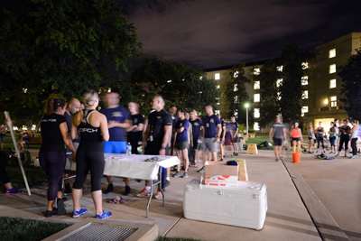 After dark in an outdoor courtyard, dozens of individuals in athletic gear line up behind a table.
