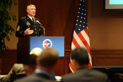 A man in a military uniform stands behind a lectern.  To his left is an American flag.  Others are seated in front of him.
