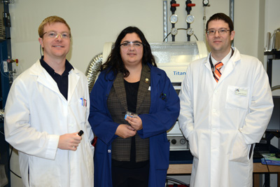 A woman in a blue lab coat is flanked by two men in white lab coats.  They stand in a laboratory.