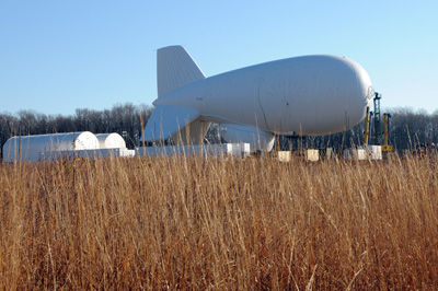 A large white blimp is attached to a mast and is situated in the middle of a field.